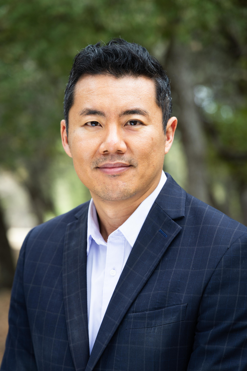 Head-and-shoulders portrait of a man with short dark hair wearing a navy blazer and light shirt, looking at the camera with a neutral expression; blurred green outdoor background.